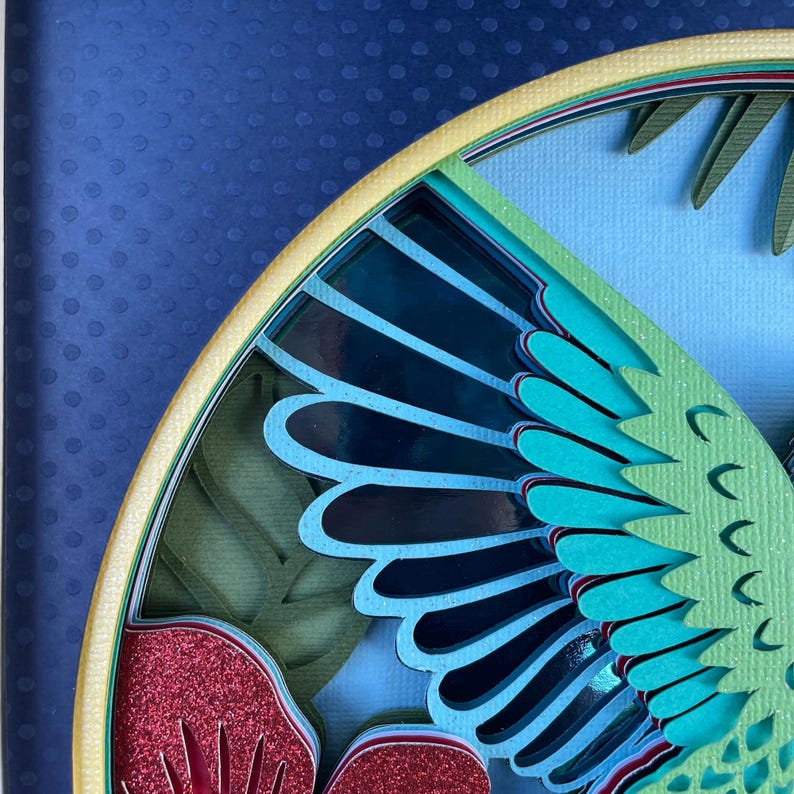 May include: Close-up of a layered paper art piece featuring a stylized bird or insect wing. The artwork uses shades of blue, teal, green, and red with glitter accents. The background is a dark blue with a dotted pattern.