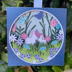 May include: A layered paper art piece featuring a spring scene with white rabbits, butterflies, and colorful flowers. The artwork is set within a circular frame, surrounded by a blue square with a dotted pattern. The scene includes green grass and stylized trees.