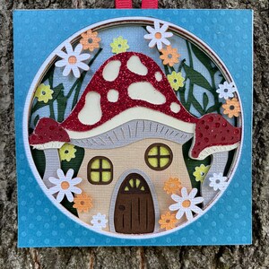 May include: A red and white mushroom with glitter accents is crafted into a whimsical house with a brown door and yellow windows. The mushroom house is surrounded by flowers and leaves, all cut from paper and layered to create a 3D effect.
