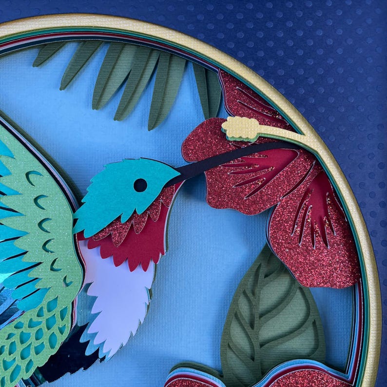 May include: A layered paper art piece featuring a hummingbird with a teal head, red throat, and white chest. The bird is positioned near a red glitter hibiscus flower and green leaves, set against a light blue background.