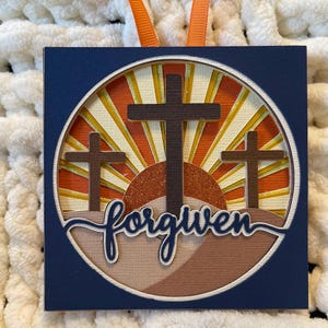 May include: A blue square ornament with a circular design featuring three brown crosses against a sunset background. The text "forgiven" is written in a cursive font at the bottom of the circle.