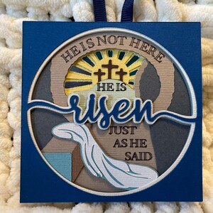 May include: A blue square with a white circle containing a layered paper design. The design features a sunrise, three crosses, and the words "He is not here, He is risen, just as He said".