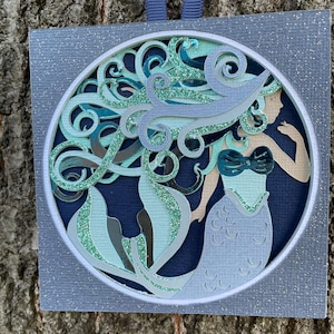 May include: A layered paper art piece featuring a mermaid design. The artwork showcases a mermaid with flowing teal hair and a blue and teal tail, set against a dark blue background within a circular frame. The frame is surrounded by a glittery, square border.