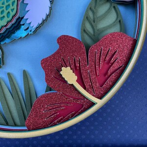 May include: A layered paper art piece featuring a red glitter hibiscus flower with a yellow stamen, green leaves, and a blue background. The artwork is circular and has a multi-layered design, creating a 3D effect.