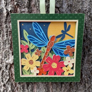 May include: A square, layered paper art piece featuring a dragonfly and flowers. The dragonfly has blue wings and an orange body, set against a yellow moon and a dark blue sky. The flowers are red, yellow, and orange, with green leaves. The frame is green.
