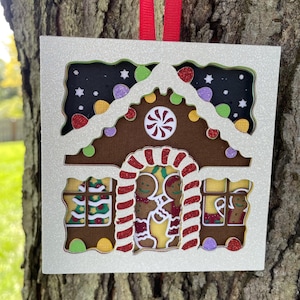 May include: A white glitter gingerbread house ornament with a red and white candy cane archway, a brown gingerbread roof, and two gingerbread people inside. The ornament has a white glitter background with a black and white starry night sky.