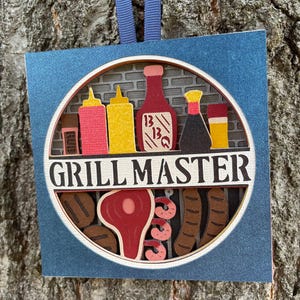 May include: A blue and white papercraft ornament with the words "Grill Master" in black lettering. The ornament features a circular design with a grill grate, various grilling condiments, and a steak.