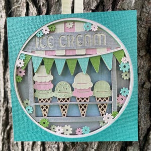 May include: A teal blue square card with a round cutout featuring a gray ice cream stand with a sign that reads "ICE CREAM". The stand has a green and blue bunting and four ice cream cones with pink and green scoops. The background is decorated with small pink and green flowers.
