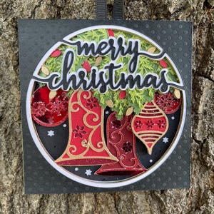 May include: A layered Christmas ornament with a black background and a circular design. The ornament features red bells, green foliage, and the words "merry christmas" in cursive. Gold and red accents add detail.