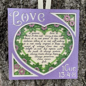 May include: A square lavender wall hanging featuring the word Love at the top and 1 Cor 13:4-8 at the bottom. A central heart frame contains the text of the passage in black script, surrounded by green die-cut leaves and small pink glitter flowers.