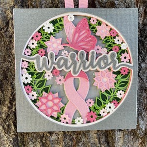 May include: A layered paper ornament with a gray background, featuring a pink ribbon, butterfly, and flowers. The word "Warrior" is prominently displayed. A pink ribbon hangs from the top.