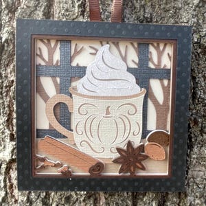May include: A framed paper art piece featuring a coffee mug with whipped cream, a pumpkin design, and fall spices. The frame is black with a dotted pattern. A brown ribbon is attached for hanging. The background shows a window with trees.