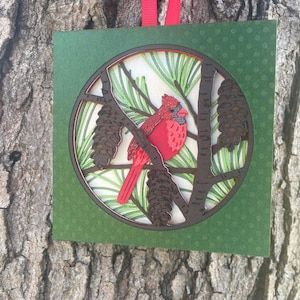 May include: A square, green paper ornament with a circular cutout featuring a red cardinal bird perched on a tree branch with pinecones. The background shows pine needles. A red ribbon is attached for hanging.