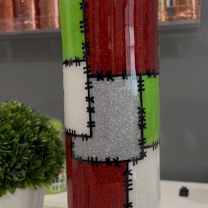 May include: A cylindrical tumbler with a Frankenstein's monster design. The tumbler is divided into sections of red glitter, green, white, and silver glitter, with black stitching details. The tumbler sits on a white base.