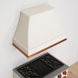 May include: White kitchen range hood with a copper trim, mounted above a black and silver stovetop.