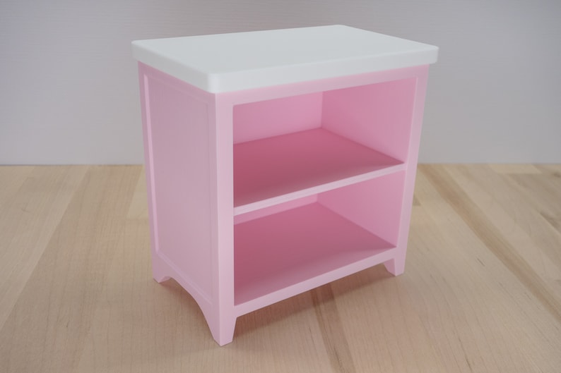 Pode incluir: Uma pequena mesa de cabeceira de madeira rosa com duas prateleiras. A parte superior &eacute; branca. A mesa tem uma forma retangular com pernas curvas. A mesa &eacute; pintada em rosa s&oacute;lido. Dimens&otilde;es: aproximadamente 50 cm x 40 cm x 30 cm.