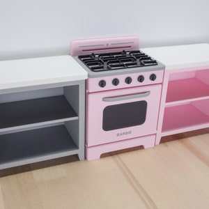 Pode incluir: Um conjunto de cozinha de brinquedo rosa com fog&atilde;o, forno e dois arm&aacute;rios laterais. O fog&atilde;o tem uma placa preta e uma porta de forno. Os arm&aacute;rios t&ecirc;m prateleiras para guardar coisas. A palavra "BARBIE" est&aacute; impressa no forno.