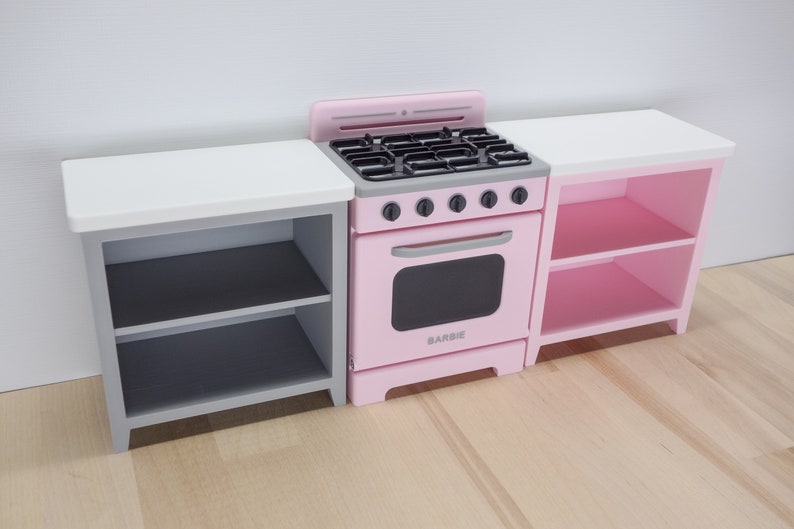 Pode incluir: Um conjunto de cozinha de brinquedo rosa com fog&atilde;o, forno e dois arm&aacute;rios laterais. O fog&atilde;o tem uma placa preta e uma porta de forno. Os arm&aacute;rios t&ecirc;m prateleiras para guardar coisas. A palavra "BARBIE" est&aacute; impressa no forno.