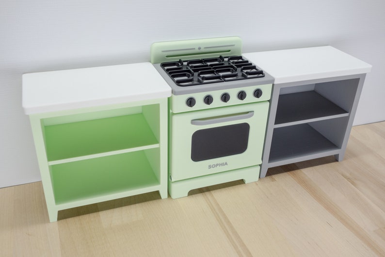 Pode incluir: Um conjunto de cozinha de brinquedo verde menta com fog&atilde;o, forno e dois arm&aacute;rios laterais. O fog&atilde;o tem uma placa preta e a palavra "SOPHIA" na porta do forno. Os arm&aacute;rios t&ecirc;m bancadas brancas e prateleiras.