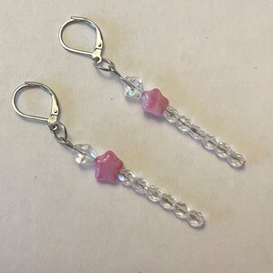 May include: A pair of dangle earrings with silver hooks and a pink star charm. The earrings are made with clear glass beads.