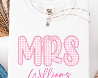 May include: White t-shirt featuring the word "MRS" in pink gingham lettering, a pink bow, and the name "Williams" in cursive. A silver necklace with a pendant is on the shirt. Blue jeans and white slip-on shoes are also visible.