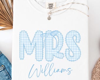 May include: White t-shirt featuring the word "MRS" in a light blue gingham design, a matching bow, and the name "Williams" in script. The image includes blue jeans and white trainers, suggesting a relaxed, everyday style.