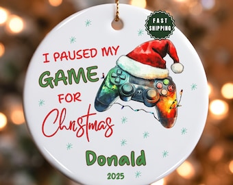 May include: A white ceramic Christmas ornament with a video game controller wearing a Santa hat. The text reads "I PAUSED MY GAME FOR Christmas" and "Donald 2025". Green and red accents with snowflake details.