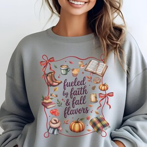 May include: A light gray crewneck sweatshirt featuring a central graphic with the text "fueled by faith & fall flavors" surrounded by illustrations of boots, a coffee mug, an open book, pumpkins, a candle, and a plaid scarf, all framed by red ribbons.