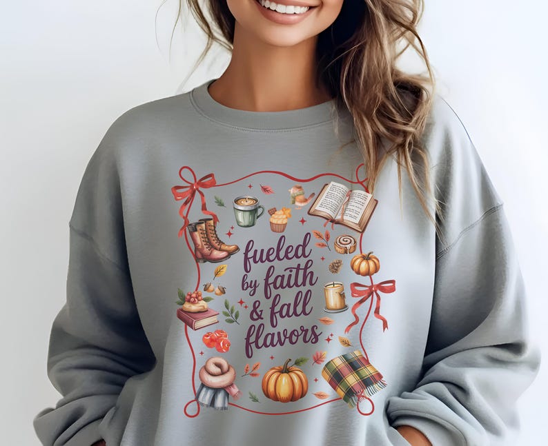 May include: A light gray crewneck sweatshirt featuring a central graphic with the text "fueled by faith & fall flavors" surrounded by illustrations of boots, a coffee mug, an open book, pumpkins, a candle, and a plaid scarf, all framed by red ribbons.