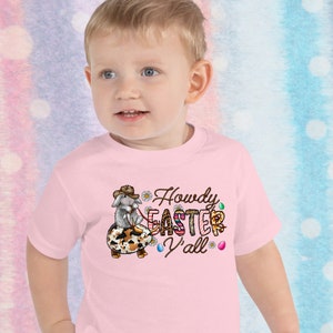 May include: A pink toddler t-shirt with a cartoon bunny wearing a cowboy hat and riding an Easter egg. The text on the shirt reads "Howdy Easter Y'all".