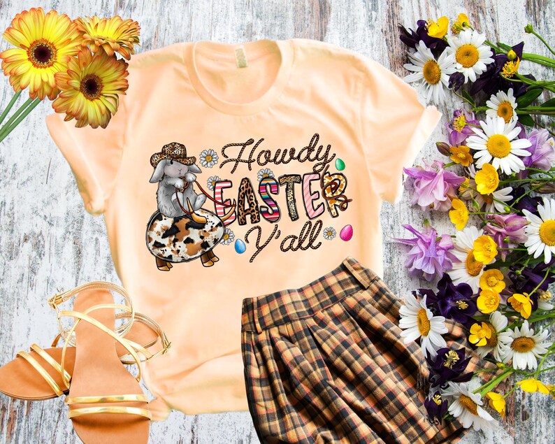 May include: A light orange t-shirt with a cartoon bunny wearing a cowboy hat and riding a decorated egg. The text "Howdy Easter Y'all" is printed on the shirt. The shirt is paired with a brown and black plaid skirt.