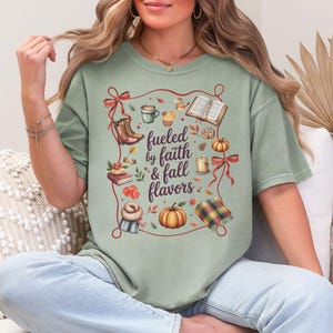 May include: A sage green t-shirt featuring a central graphic with the text "fueled by faith & fall flavors" surrounded by illustrations of boots, a book, a pumpkin, a candle, a scarf, a plaid blanket, and red ribbons on a light background.