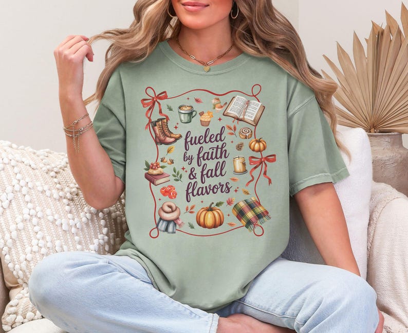 May include: A sage green t-shirt featuring a central graphic with the text "fueled by faith & fall flavors" surrounded by illustrations of boots, a book, a pumpkin, a candle, a scarf, a plaid blanket, and red ribbons on a light background.