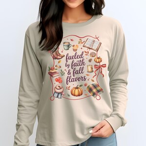 May include: A beige long-sleeve shirt featuring a central graphic with the text "fueled by faith & fall flavors" surrounded by illustrations of a coffee mug, an open book, a pumpkin, a candle, a plaid blanket, boots, and autumn leaves.