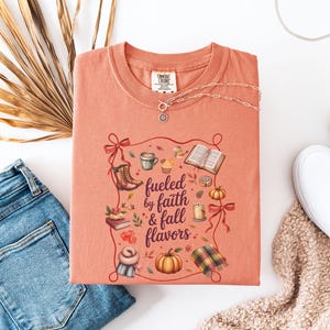 May include: A coral-colored crewneck t-shirt featuring a graphic print with the text "fueled by faith & fall flavors" surrounded by illustrations of boots, a coffee mug, an open book, pumpkins, a candle, a scarf, and a plaid blanket.