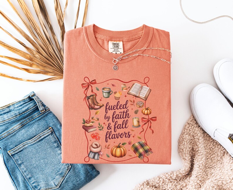 May include: A coral-colored crewneck t-shirt featuring a graphic print with the text "fueled by faith & fall flavors" surrounded by illustrations of boots, a coffee mug, an open book, pumpkins, a candle, a scarf, and a plaid blanket.