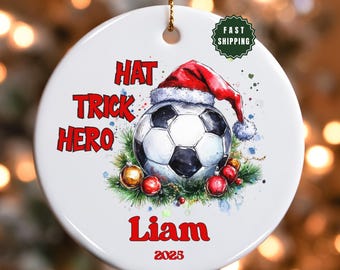 Op de afbeelding: Witte keramische kerstbal met een voetbal met een kerstmuts. De bal heeft de woorden "HAT TRICK HERO" en de naam "Liam" in het rood. Inclusief een "FAST SHIPPING" sticker.