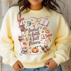 May include: A pale yellow crewneck sweatshirt featuring a central graphic with the text "fueled by faith & fall flavors" surrounded by illustrations of boots, a coffee mug, an open book, a pumpkin, a candle, a scarf, and autumn leaves.
