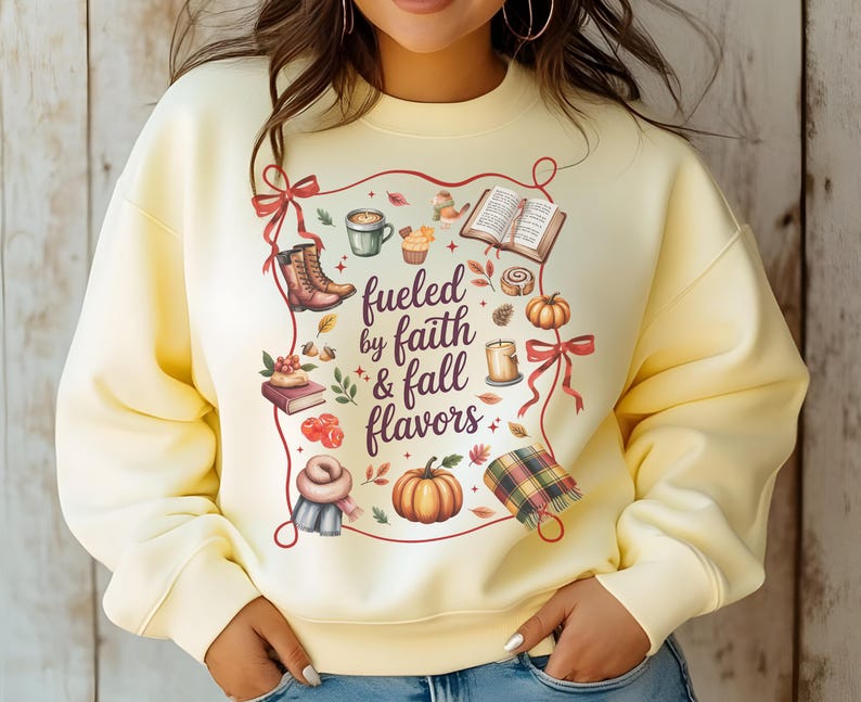May include: A pale yellow crewneck sweatshirt featuring a central graphic with the text "fueled by faith & fall flavors" surrounded by illustrations of boots, a coffee mug, an open book, a pumpkin, a candle, a scarf, and autumn leaves.