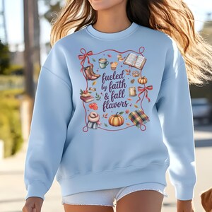 May include: A light blue crewneck sweatshirt featuring a central graphic with the text "fueled by faith & fall flavors" surrounded by illustrations of a coffee mug, an open book, a pumpkin, a candle, boots, a scarf, and red ribbon accents.