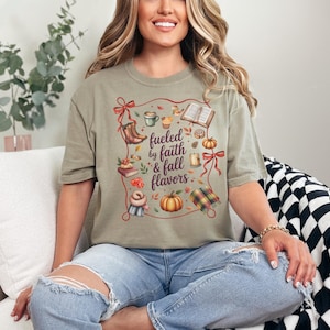 May include: A sage green t-shirt featuring a graphic print with the text "fueled by faith & fall flavors" surrounded by illustrations of a pumpkin, a mug, books, boots, a scarf, a candle, and a plaid blanket, all framed by a red ribbon border.