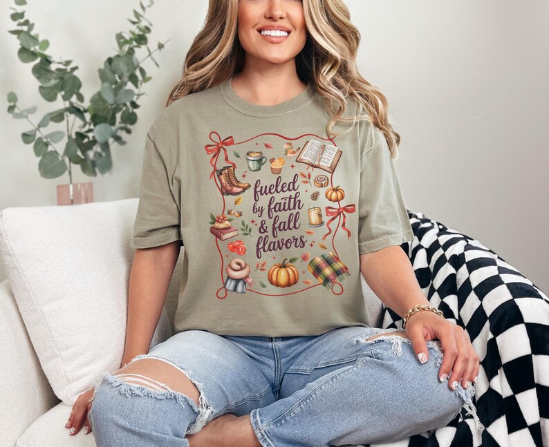 May include: A sage green t-shirt featuring a graphic print with the text "fueled by faith & fall flavors" surrounded by illustrations of a pumpkin, a mug, books, boots, a scarf, a candle, and a plaid blanket, all framed by a red ribbon border.