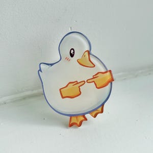 May include: A clear acrylic duck-shaped pin with a blue outline. The duck has a white body, orange beak and feet, and orange hands pointing at itself. The duck has a blushing face.