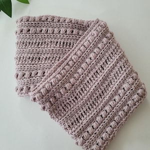 Può includere: Un caldo collo in crochet rosa con un motivo a punto testurizzato. Il collo è piegato a metà e ha un bordo leggermente arricciato.