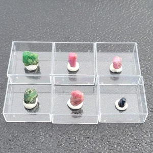 Puede incluir: Seis pequeñas cajas de plástico transparente que contienen cada una una gema de color diferente. Las gemas son verdes, rosas, rojas y azules.