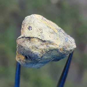 Puede incluir: Primer plano de una roca sujeta con pinzas. La roca tiene una superficie rugosa y texturizada con una mezcla de colores amarillo, gris y marrón. La forma de la roca es irregular, con grietas visibles y variaciones de color.