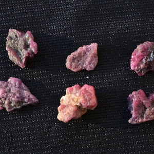 May include: Six rough, pink ruby gemstones on a black fabric background.