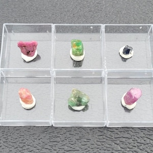 Puede incluir: Seis pequeñas cajas de plástico transparente que contienen cada una una gema diferente. Las gemas son un rubí rosa, una esmeralda verde, un zafiro negro, un rubí rosa, una esmeralda verde y un rubí rosa.