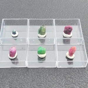 May include: Six small, colourful gemstones displayed in clear plastic boxes. The gemstones are various shades of green, pink, and blue.