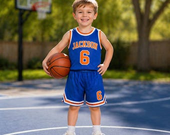 May include: A young child wearing a blue basketball jersey and shorts with orange trim, the name "JACKSON" and the number "6" are visible. The child is holding a basketball and smiling. The background includes a basketball hoop and trees.